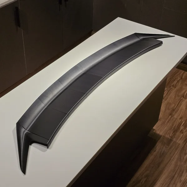 Black 3D printed carbon fiber car spoiler wing displayed on a counter by Print3D.pk.